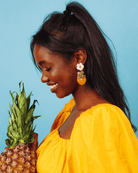 all-groups Femme métisse souriant portant un ananas. elle est habillée avec un haut jaune et des boucles d'oreilles fruits jaunes.  