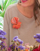 all-groups Femme brune souriant vetue d'un pull scintillant beige sur lequel est accroché une broche fleur orange